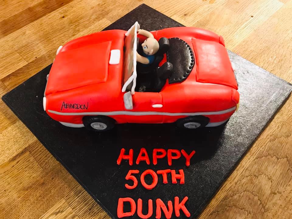 A cake shaped into a sports car covered in red fondant with a fondant figure sitting in the drivers seat