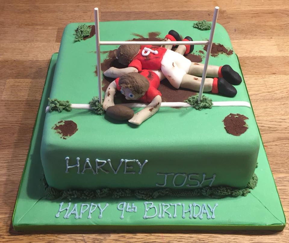 Square cake covered in green fondant with 2 fondant Ruby players on top in a scrum