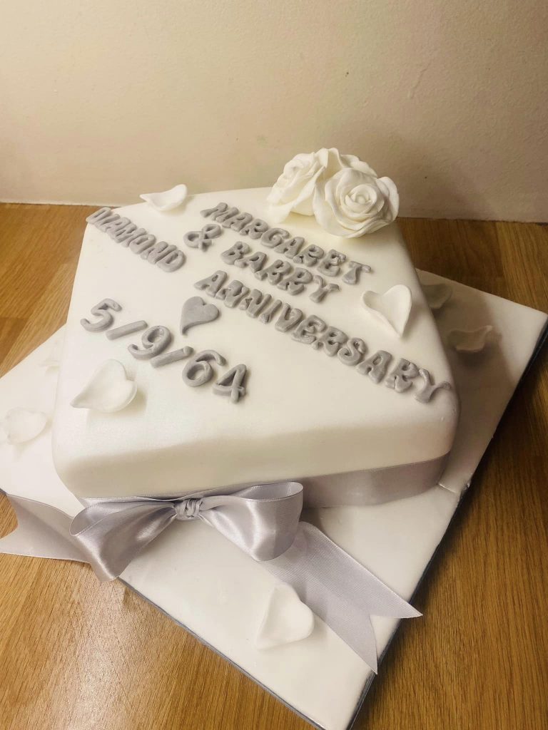 Diamond shaped cake covered in white fondant with silver wording, fondant hearts and a ribbon around the cake by creative cakes by kaz