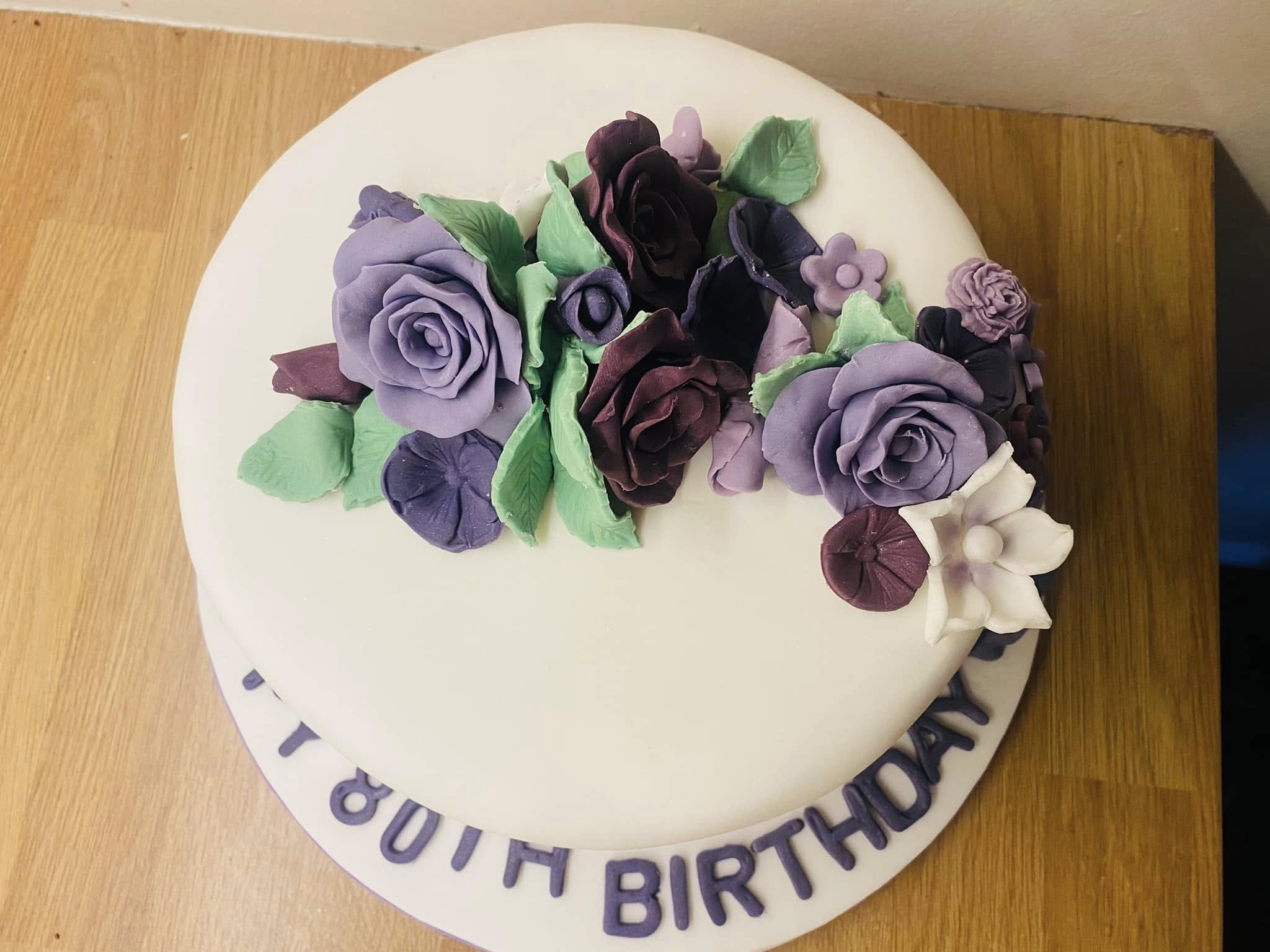 Round cake covered in white fondant with lilac and purple fondant flowers on top 