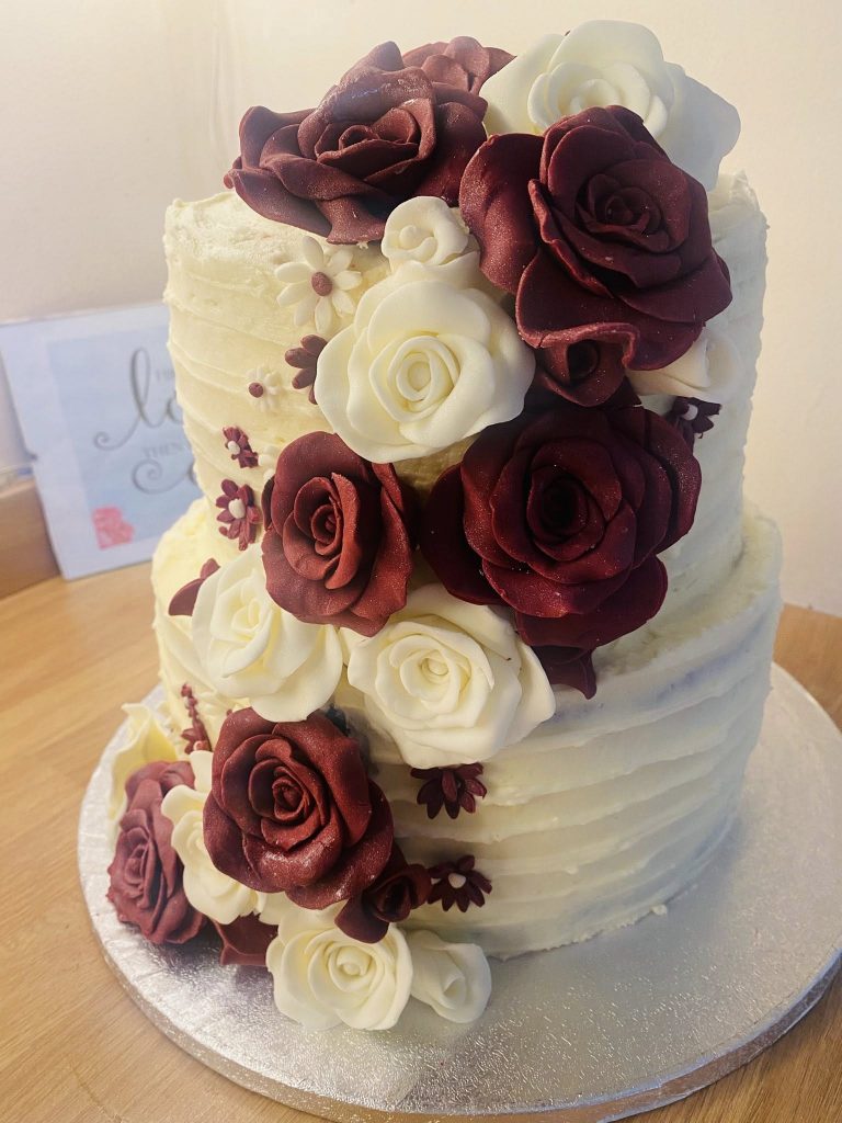 2 tier wedding cake covered in cream buttercream with cream and burgundy fondant roses trailing down cake by Creative Cakes by Kaz