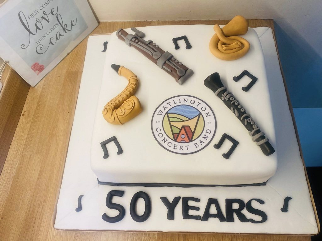 Square cake covered in white fondant with the Watlington Concert Band logo and fondant musical items on top 
