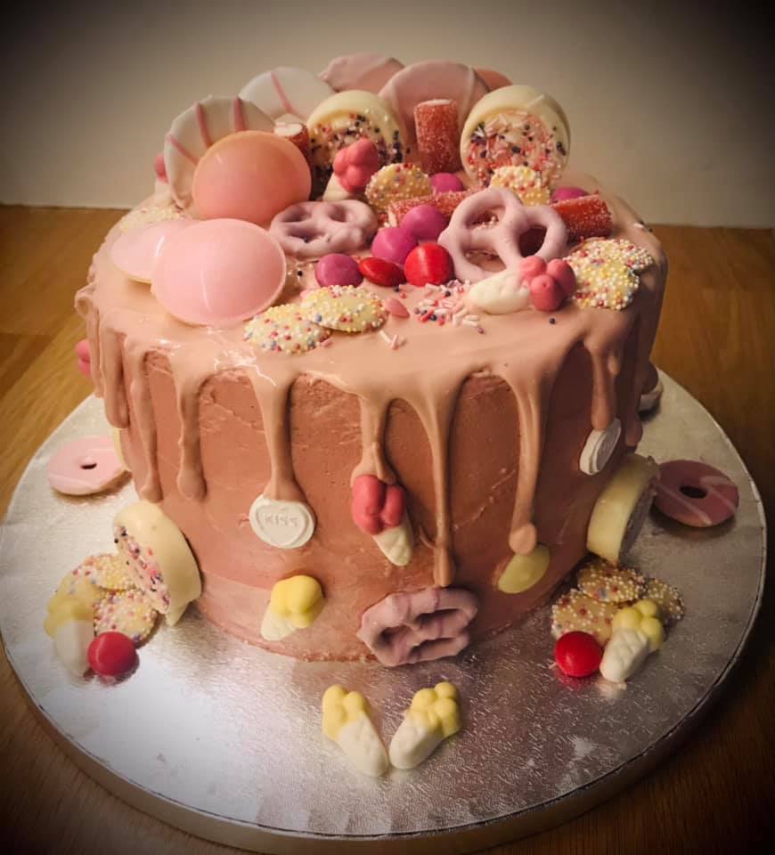 Round cake covered in pink buttercream with a pink chocolate drip with lots of different sweets on top 